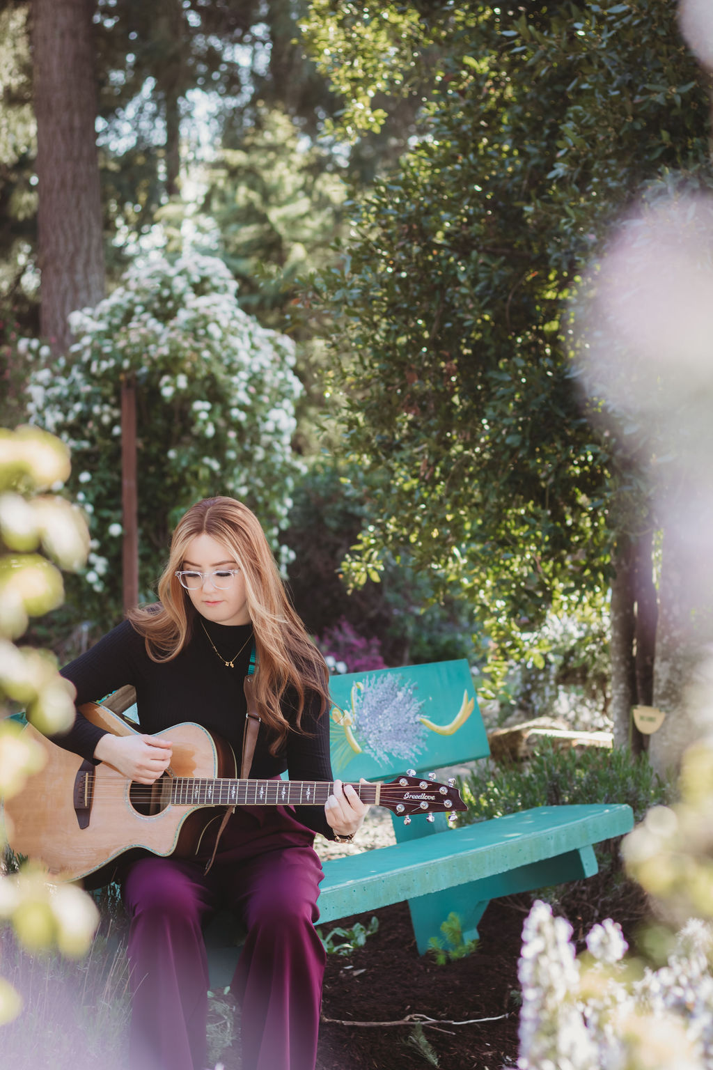 A woman plays guitar during a branding photography session at Discovery Garden in Roseburg, Oregon, surrounded by spring greenery and soft sunlight. Captured by McKenzie Strong Photography for a creative headshot and branding session.