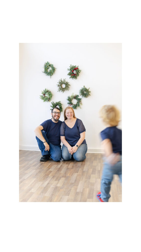 Parents photographed during a Roseburg Oregon Christmas mini session while their toddler runs toward them in a bright studio with holiday wreaths